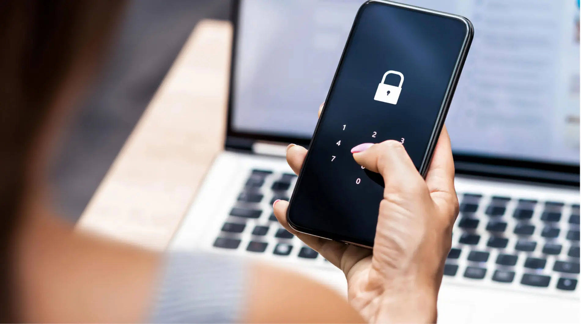 Smartphone con lucchetto sullo schermo, tenuto in mano. Sullo sfondo, un laptop.