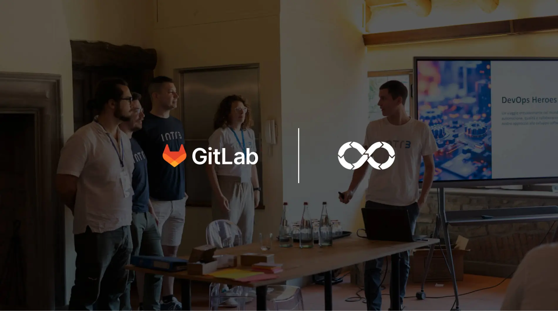Foto con sullo sfondo persone che parlano, e in primo paino loghi di GitLab e doppio loop DevOps