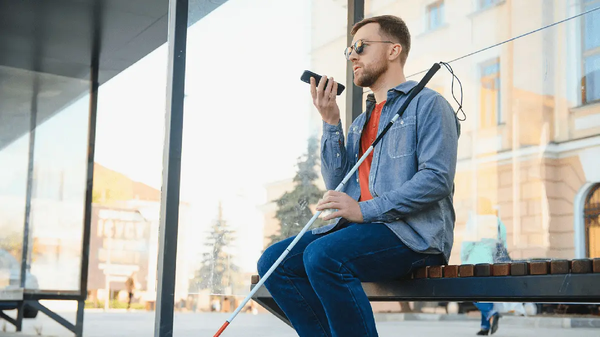 Di giorno, un uomo con disabilità è seduto alla fermata del bus e sta mandando un audio con il telefono
